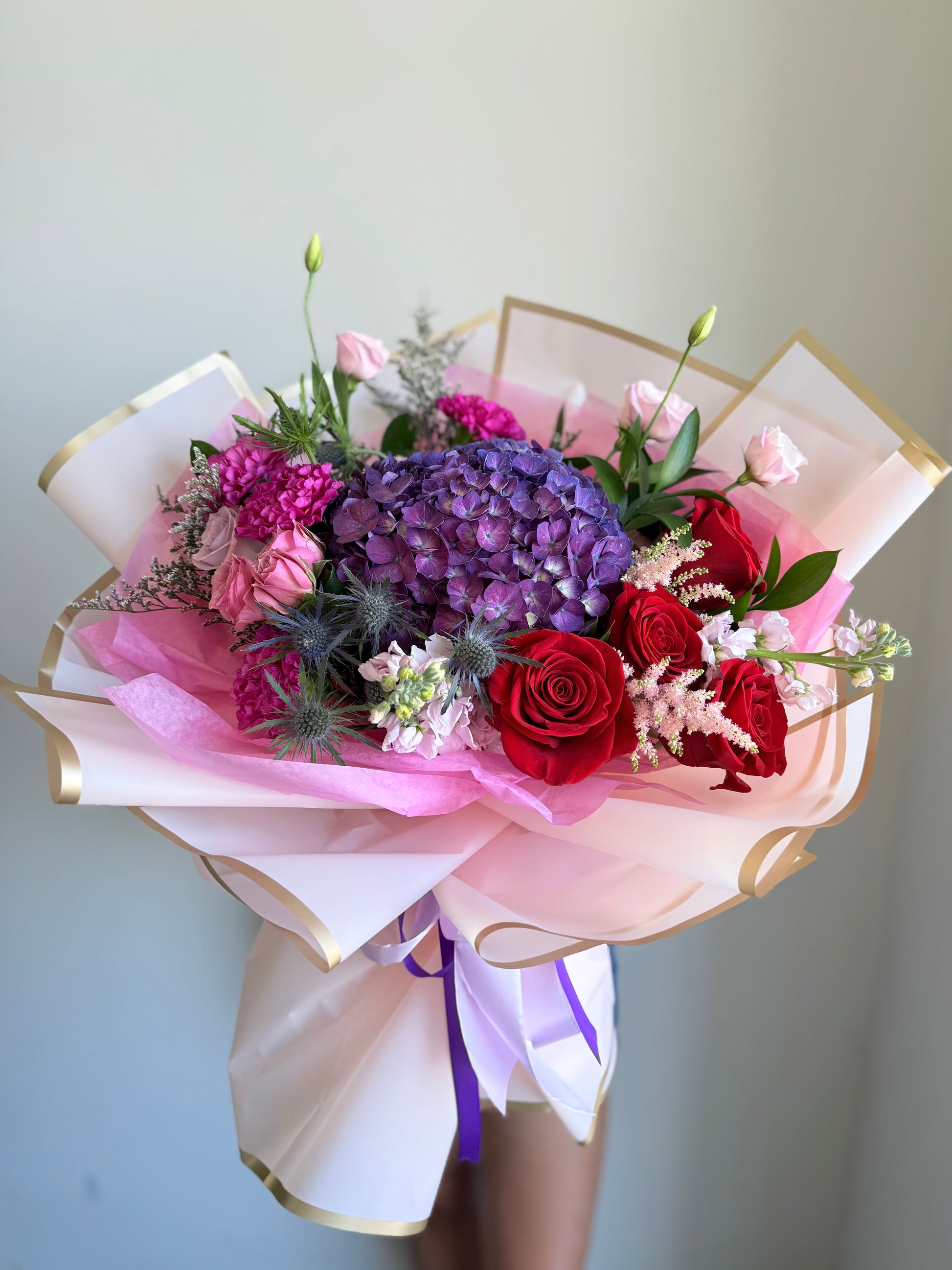 Deeply Yours Bouquet of flowers with red roses, purple hydrangeas, and pink flowers in a white and gold wrap on a light gray background.